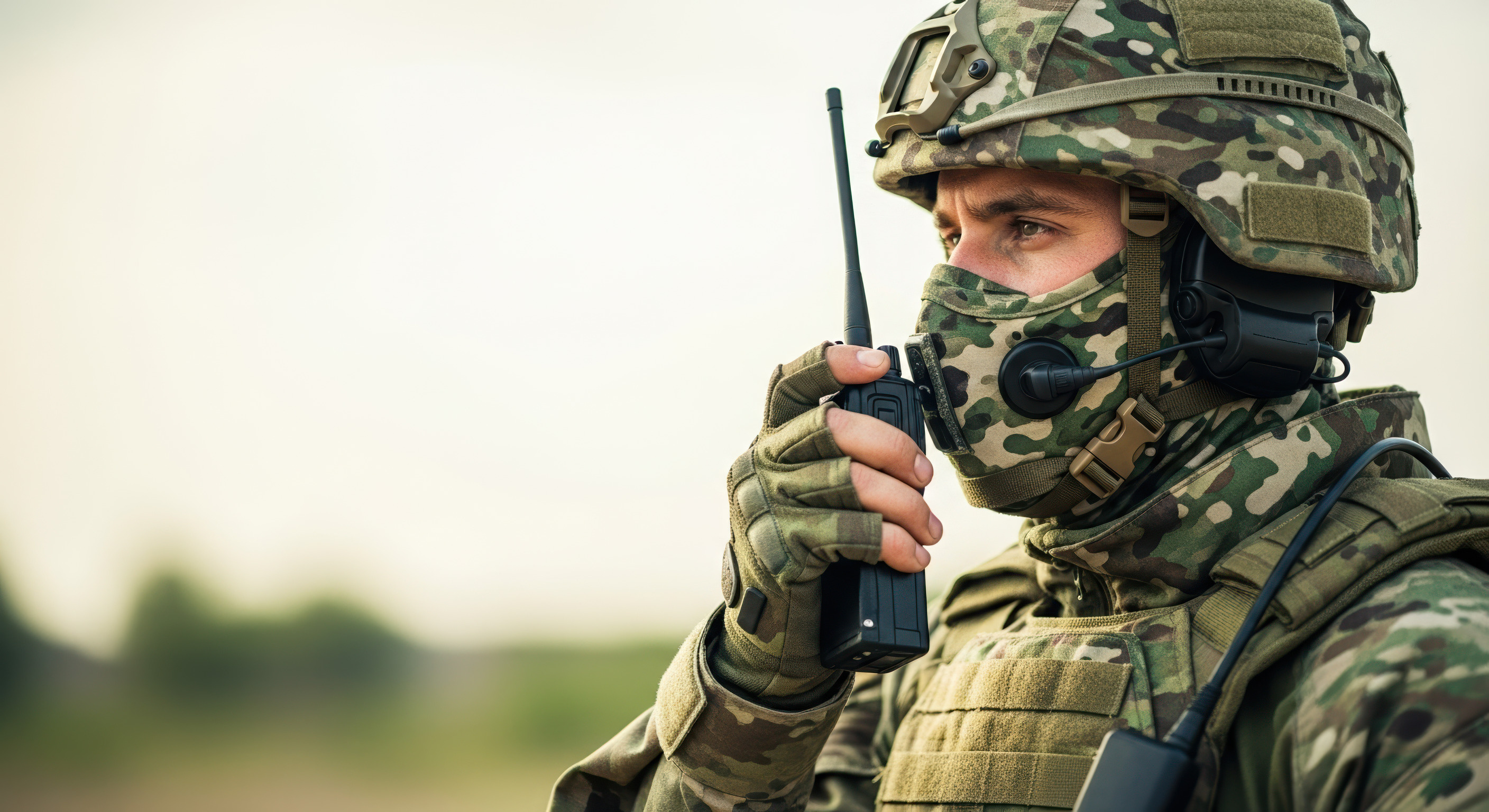 A soldier in camouflage helmet and face covering speaks into a handheld tactical radio, headset fitted, with soft open sky to the left.