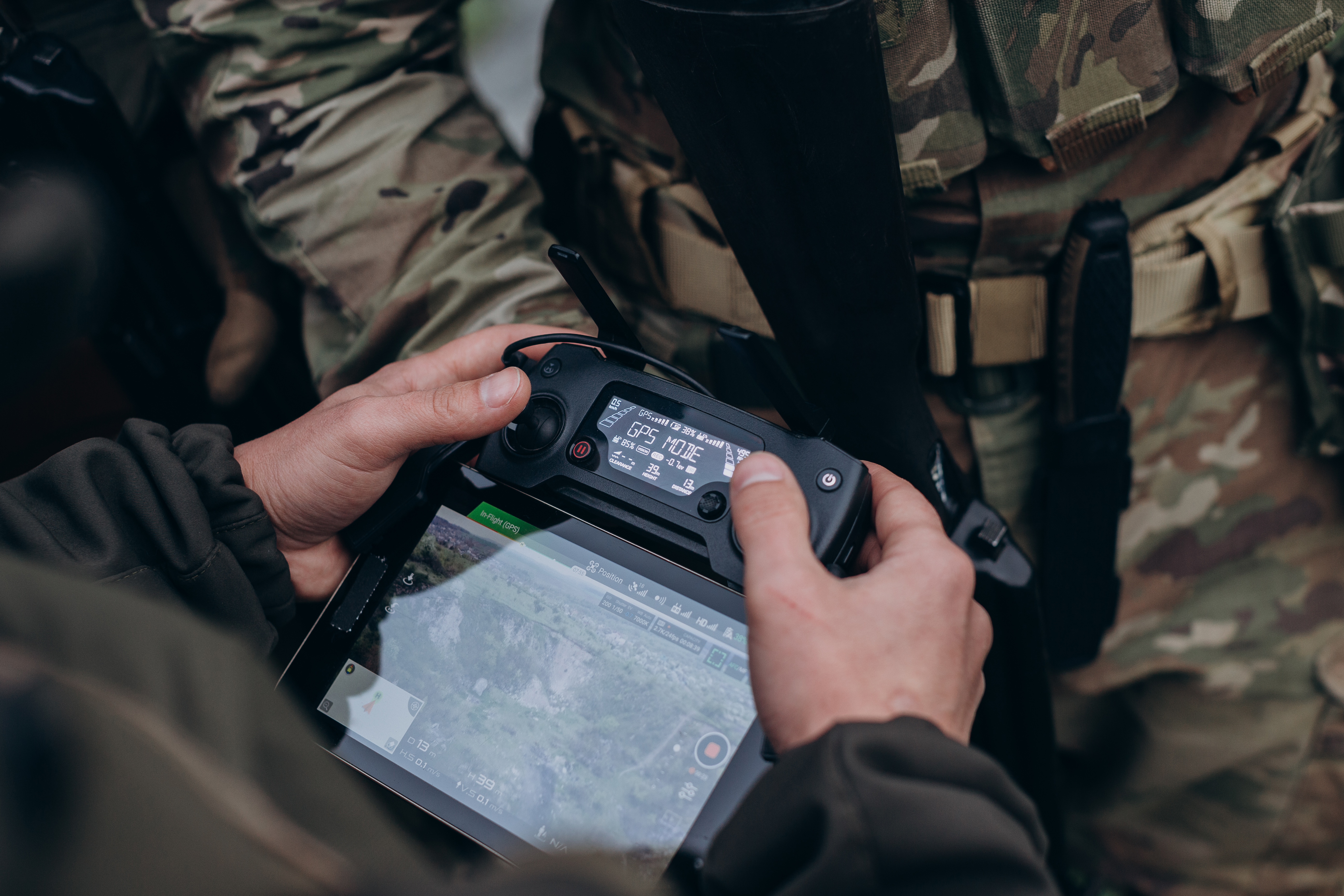Overhead view of gloved hands operating a drone controller with a lit telemetry display reading GPS mode, above a tablet showing an aerial map, against camouflage clothing.